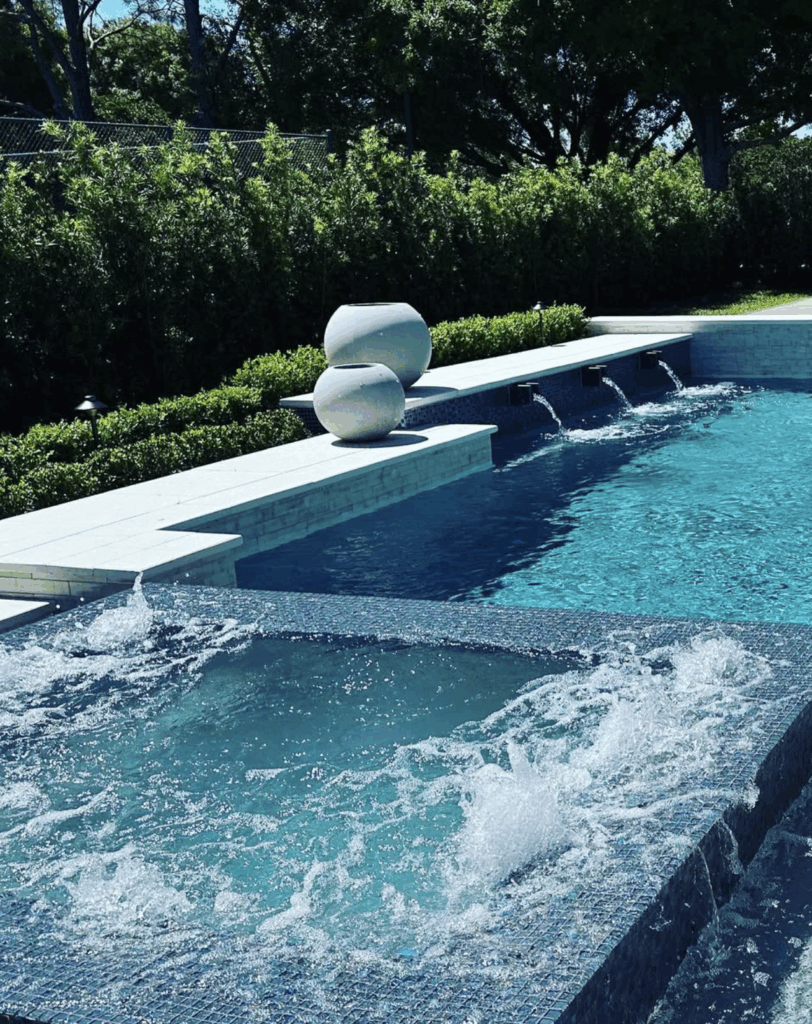 A modern pool with a bubbling hot tub in the foreground, three small waterfalls, and decorative white vases along the pool edge surrounded by greenery.